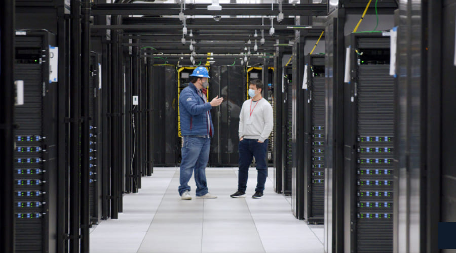Two technicians wearing face masks and a hard hat stand in a data center aisle discussing maintenance among rows of server racks.