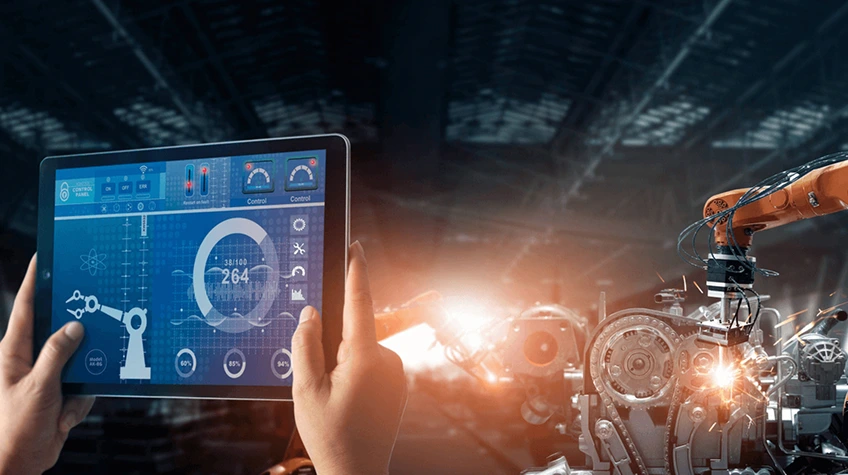 A first-person view of a person holding a tablet that displays a real-time diagnostic dashboard. In the background, a robotic arm performs precision welding on an automotive assembly line.