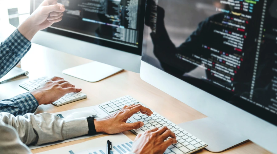 Two people working at a desk with large computer monitors displaying lines of code.