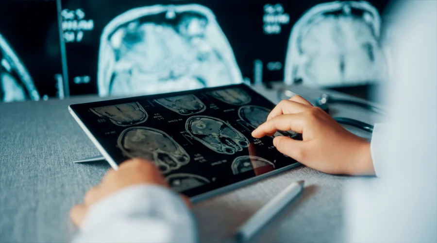 A healthcare professional using a tablet to examine detailed brain MRI scans, with larger monitors in the background.