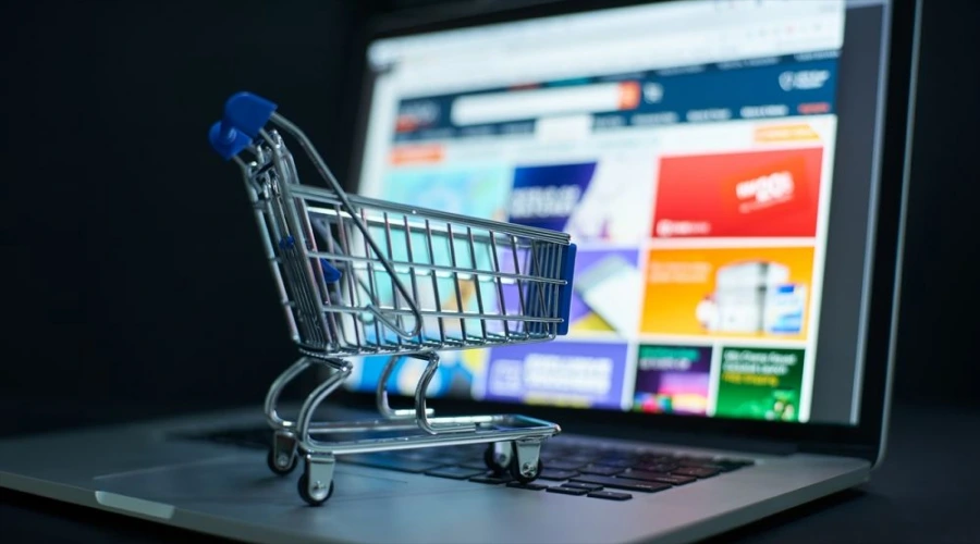 A miniature shopping cart sitting on a laptop keyboard in front of a blurred online retail website.