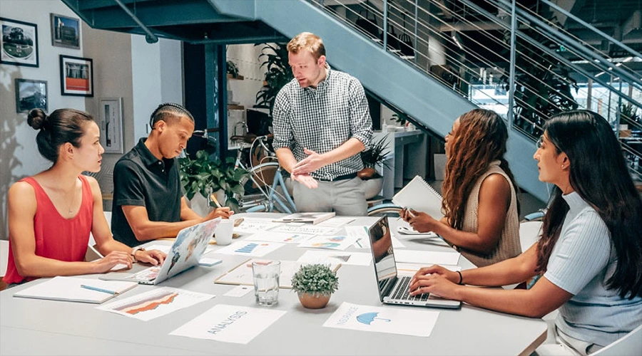 Better Collaboration Between Teams - WeeTech Solution Pvt Ltd A diverse team discussing documents and working on laptops around an office conference table.
