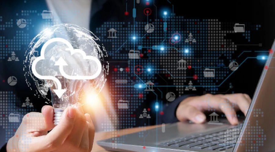 A hand holding a glowing lightbulb with a cloud icon inside, next to a person typing on a laptop with digital network overlays.