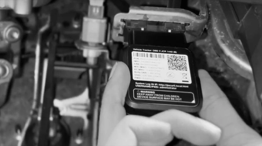 JioMotive Device How Does It Work_ - WeeTech Solution Pvt Ltd A close-up, black-and-white image of hands installing a black OBD tracker device into a vehicle's under-dashboard port.
