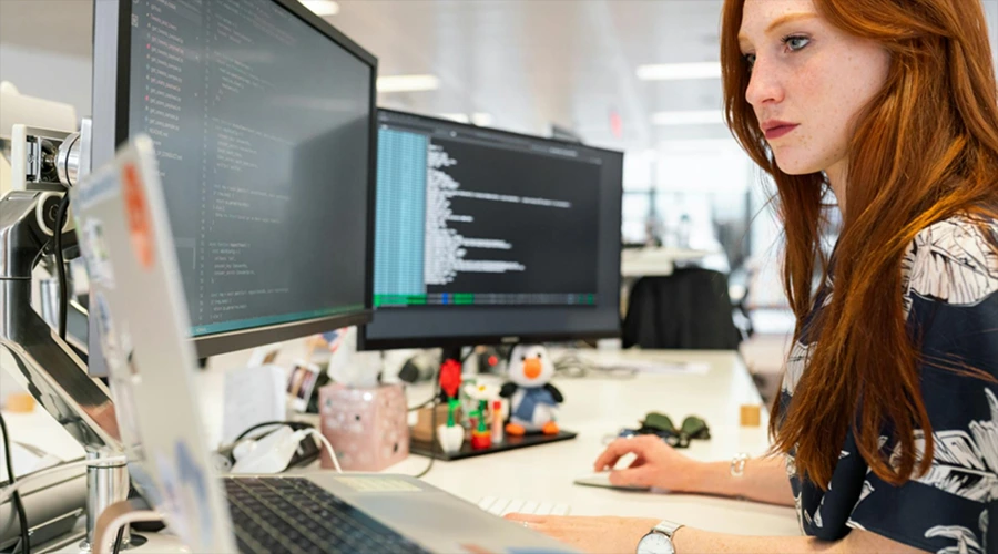Significant Reduction in Human Errors - WeeTech Solution Pvt Ltd A woman focused on reviewing code displayed on dual monitors at her desk.