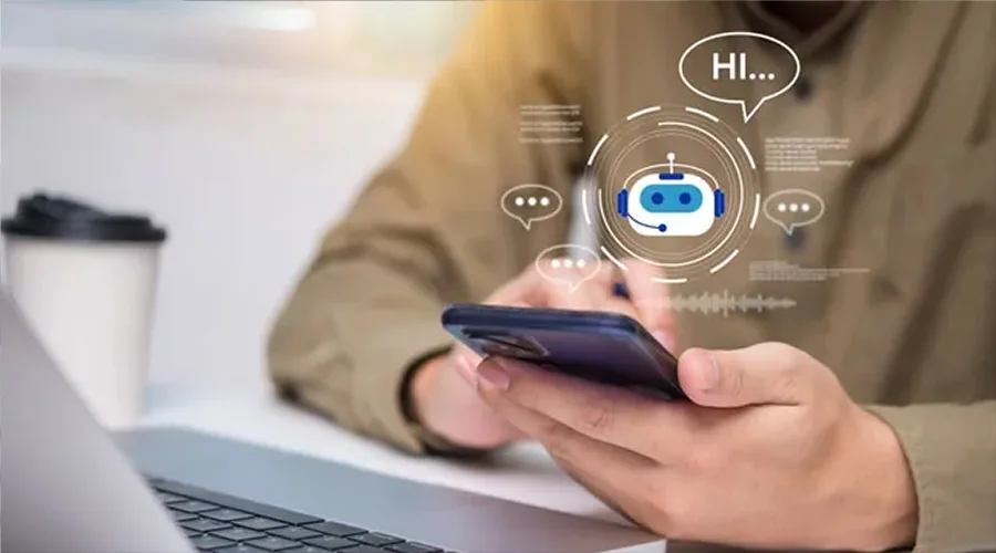 A person holding a smartphone showing an interactive AI chatbot speech bubble overlay with a laptop nearby.