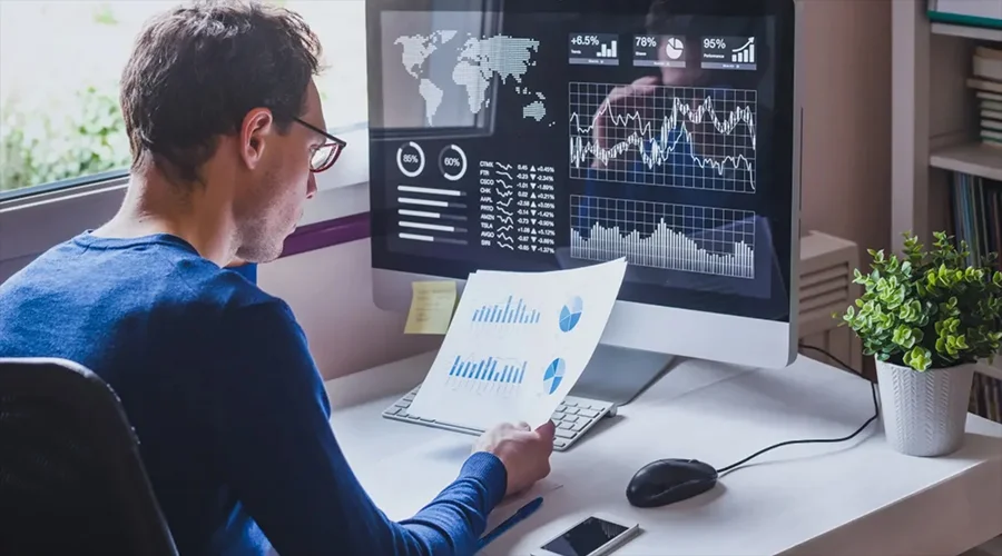 Man sitting at a desk analyzing paper reports and charts displayed on a large computer screen.