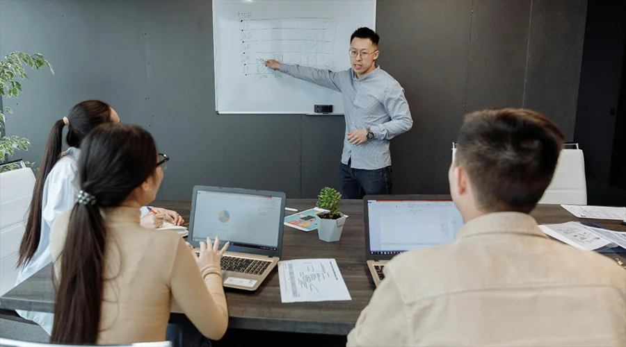 Presenter standing at a whiteboard pointing to drawn bar charts during a team meeting.